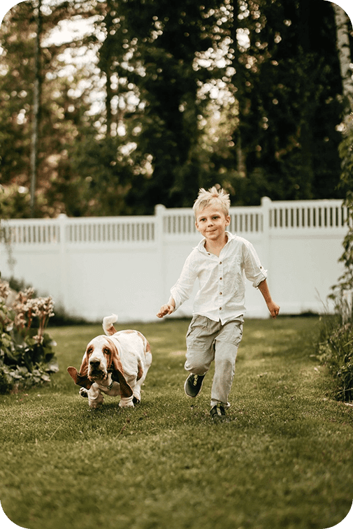 Lapsi ja koira juoksevat omakotitalon pihan halki, taustalla näkyy valkoinen vinyyliaita